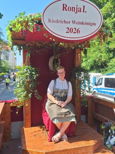Auf einem LKW ist aus Holz ein Thron gebaut, der mit Efeu, Weinblättern und Reben und rotem Stoff geschmückt ist. Auf dem Thron sitzt eine blonde Frau mit Brille, weißer Bluse und grünem Dirndl. Sie hat eine goldene Krone auf dem Kopf und ein gefülltes Weinglas in der Hand. Neben dem LKW parkt ein Polizeitransporter und stehen Dixitoiletten.