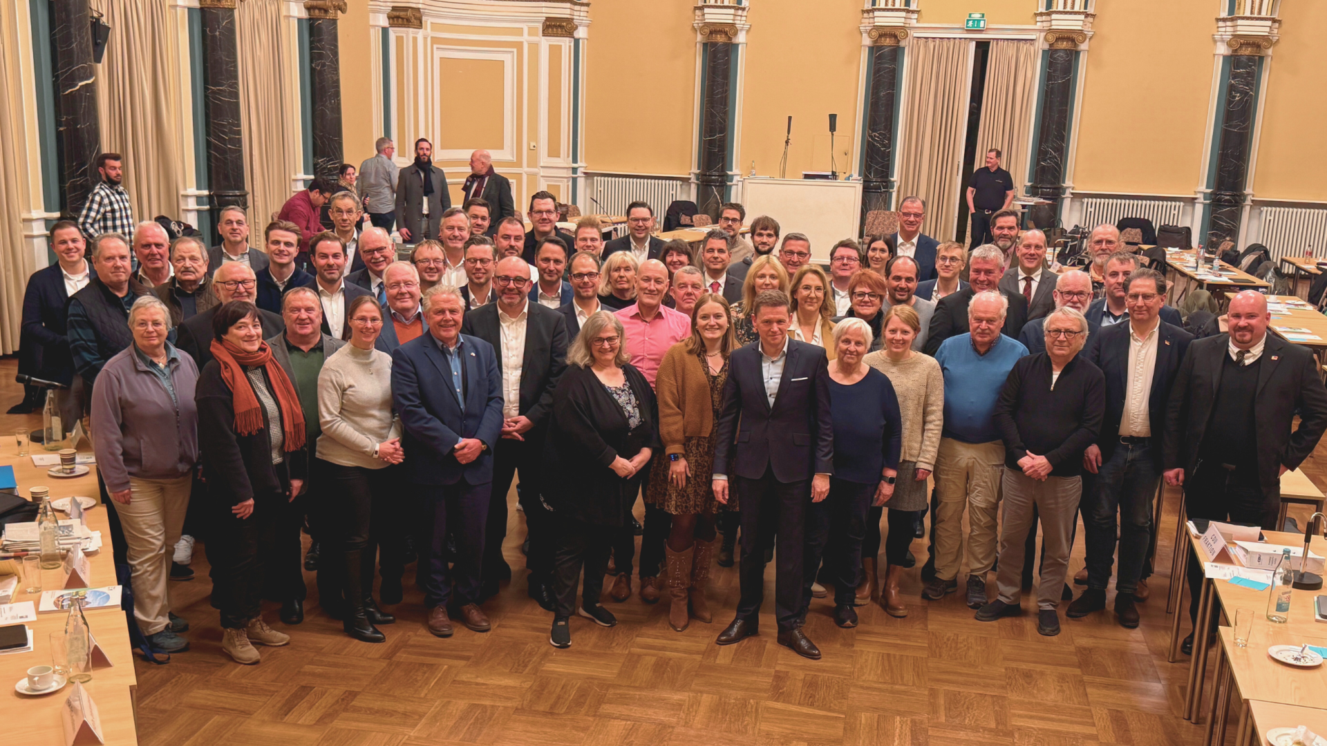 In einem großem Saal mit gelben Wänden und schwarzen Säulen mit goldenen Verzierungen und weißem Stuck, stehen über 60 Männer und Frauen. 