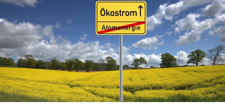 Ein gelb blühendes Rapsfeld mit grünen Laubbäumen im Hintergrund und einem blauen Himmel mit weißen Wolken. Vorne im Bild ist ein gelbes Ortsschild unten steht der Begriff Atomenergie, der rot durchgestrichen ist und oben wird Ökostrom genannt und ein schwarzer Pfeil zeigt nach oben.