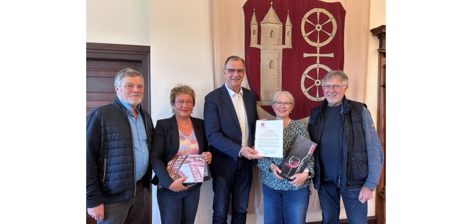 Vor dem Gemeinde-Wappen in weinrot, mit beigem Doppelrad und der Burg stehen drei Männer und zwischen den Männern zwei Frauen. Die linke Frau hat ein Heft über eine Kirche in der Hand und die andere Frau trägt ein Weinpräsent. Der Mann in der Mitte hat eine Urkunde in der Hand.