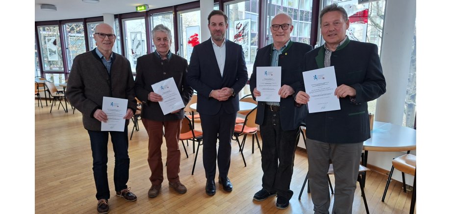 Vor einer Fensterfront und mehreren runden Tischen mit Stühlen, stehen fünf Männer in dunkler Kleidung und die jeweils zwei äußeren Personen halten weiße Urkunden in den Händen. 