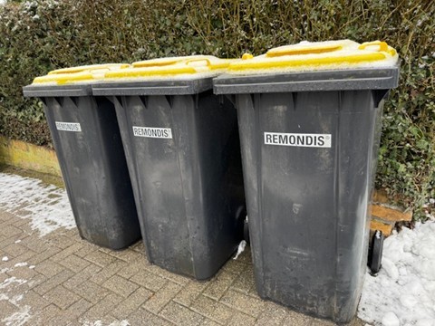 Vort einer Hecke stehen drei schwarze Tonnen mit der Aufschrift Remondis und gelben Deckeln. Auf dem Gehweg liegt zusammengeschobener Schnee. 