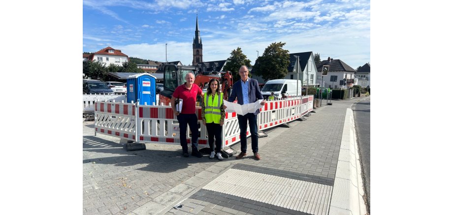 Eine Frau mit langen braunen Haaren und Warnweste steht zwischen zwei Männern, die eine Akte und einen Bauplan in den Händen haben. Sie stehen vor weiß-roten Absperrungen, hinter denen eine blaue Dixitoilette und geparkte Autos zu sehen sind. Im Hintergrund ist ein Kirchturm.