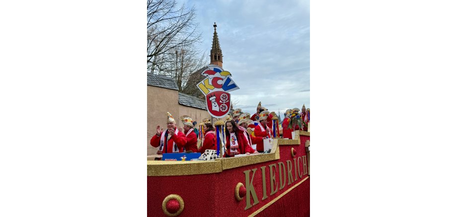 Auf dem mit rot-goldenem-Glitzerstoff geschmückten Komiteewagen sind die Großbuchstaben KIEDRICH in gold angebracht. Auf dem Wagen stehen die Vereinsmitglieder mit roten Jacken, weißen Wollschals mit dem gelb-rot-blau-weißem KCV-Logo mit dem Kiedricher Wappen, welches ebenso als Schild auf dem Waagen steht. Im Hintergrund ist der Turm der Michaelskapelle und die Kirchhofmauer und Bäume ohne Blätter zu erkennen.