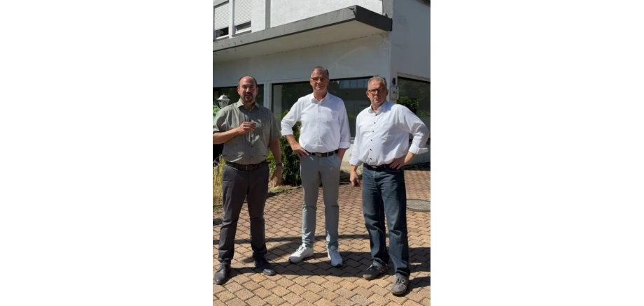 Drei Männer stehen vor einem weißem Gebäude. Zwei tragen weiße Hemden und einer ein olivfarbenes Hemd nd jeweils dunkel Hosen.