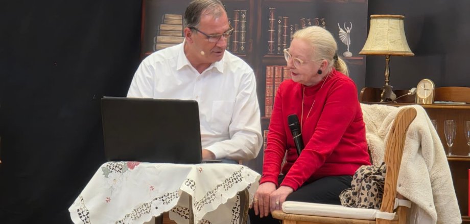 Ein Mann und eine Frau sitzen auf Holzstühlen um einen kleinen Tisch herum. Auf dem Tisch ist eine weiße Tischdecke und ein aufgeklappter Laptop. Im Hintergrund ist ein Bild eines Bücherregals und ein halbhohes Regal, in dem unter anderem eine Uhr, eine Lampe und Sektgläser stehen.