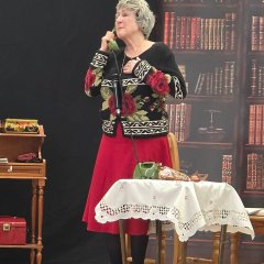 Eine Frau steht mit rotem Rock, schwarz weißer Strickjacke, mit roten Rosen und grünen Blättern und verschieden farbigen Wollsocken und grauen Hausschuhen, neben einem kleinen Tisch auf dem eine weiße Tischdecke und ein grünes Telefon steht. Den Telefonhörer hat sie in der Hand am Ohr. Man sieht die Telefonschnur. Im Hintergrund ist ein Bild eines Bücherregals und ein halbhohes Regal, in dem unter anderem Geldbörsen und Schmuckboxen liegen.