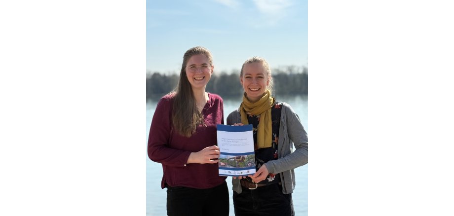 Zwei junge Frauen halten ein Heft in der Hand. Eine Frau hat lange blonde Haare, trägt eine Brille, ein weinrotes Langarm-Shirt und eine schwarze Hose. Die Frau daneben trägt eine schwarze Hose, eine schwarze Bluse mit Blumen, einer grauen, offenen Strickjacke und einen gelben Schal. Die Haare sind zu einem Zopf gebunden. Der Hintergrund ist unscharf und es ist Wasser, eine Baumreihe und Himmel mit Wolken zu erahnen.