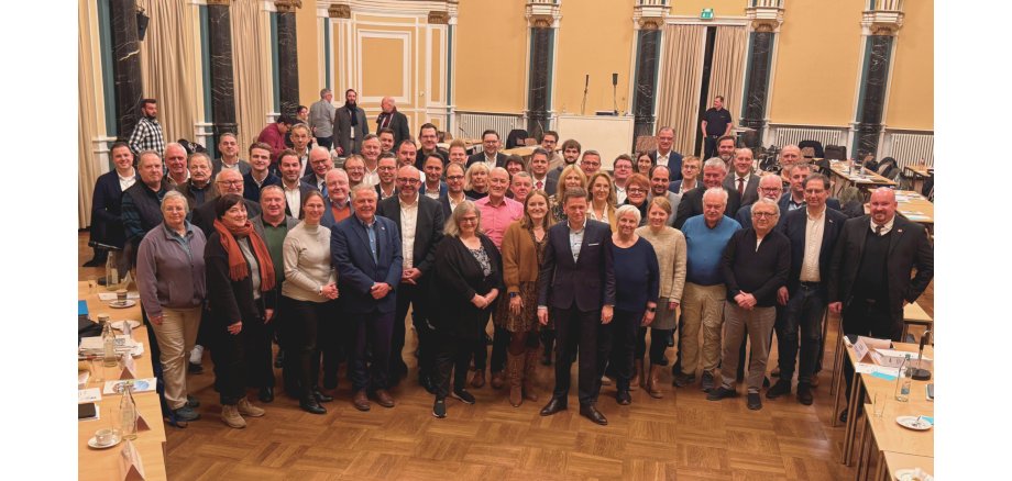 Rechtsanspruch - 2 In einem großem Saal mit gelben Wänden und schwarzen Säulen mit goldenen Verzierungen und weißem Stuck, stehen über 60 Männer und Frauen.