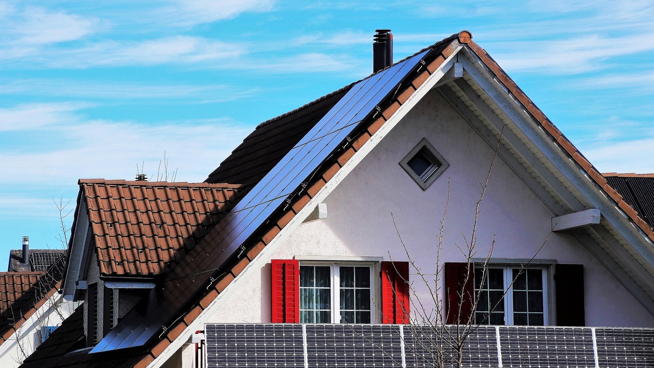 Man sieht das Dachgeschoss mit einem Dachgiebel. Hinter dem Balkon mit fünf Solarpanelen sind zwei Paar Sprossenfenster mit roten Rolläden. Das Haus ist weiß und die Dachziegel rot-braun. Auf dem Dach sind ebenso Solarpanelen.