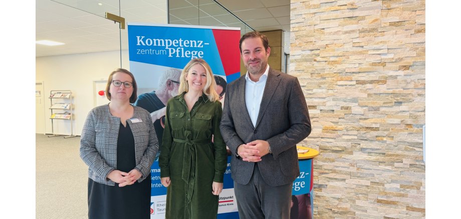 Vor einem Plaket mit der Aufschrift "Kompetenzzentrum Pflege stehen zwei Frauen und ein Mann. Die Frau in der Mitte hat blonde, lange Haare und ein grünes langes Kleid und die Frau daneben braune kinnlange Haare und eine Brille und ein schwarzes Kleid mit einer schwarz-weiß karrierten Jacke. Der MAnn trägt ein weißes Hemd und eine sunkelgraue Hose und Jacket. Rechts ist eine Steinwand zu sehen und links ein Flur mit einem Plakatständer.    