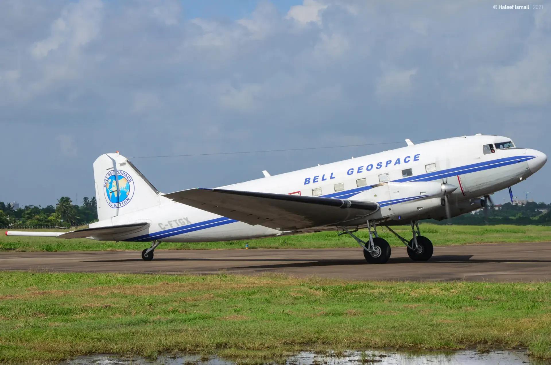 Ein kleines Flugzeug mit zwei Triebwerken steht auf einem Rollfeld. Man sieht ebenso Wiese und einen bewölkten Himmel. Das Flugzeug ist weiß mit einem blauem Streifen und blauer Schrift und einem blauem Kreis mit gelb-grünem Inhalt.