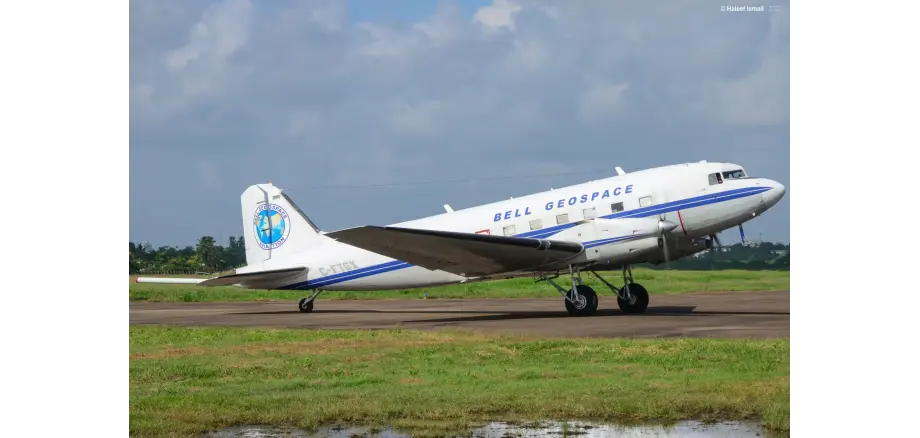 Ein kleines Flugzeug mit zwei Triebwerken steht auf einem Rollfeld. Man sieht ebenso Wiese und einen bewölkten Himmel. Das Flugzeug ist weiß mit einem blauem Streifen und blauer Schrift und einem blauem Kreis mit gelb-grünem Inhalt.