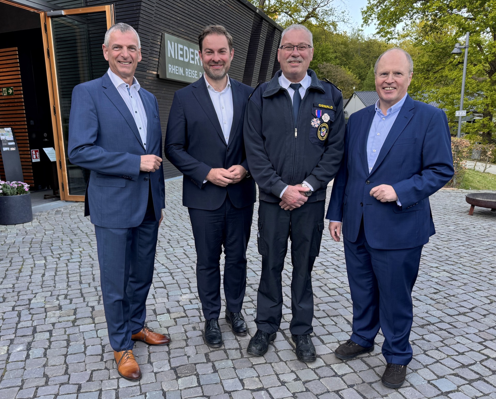 Vier Männer in hellblauem Hemd und dunkelblauem Anzug stehen vor einer Halle, die mit horizontalen Holzbalken verkleidet ist. Der zweite Mann von rechts trägt eine Krawatte, eine Brille und eine Jacke mit seinem Namen und zwei Orden sowie einer Plakette. Der Boden, auf dem sie stehen ist mit Kopfsteinpflaster gepflastert. Im Hintergrund sind grüne Bäume.