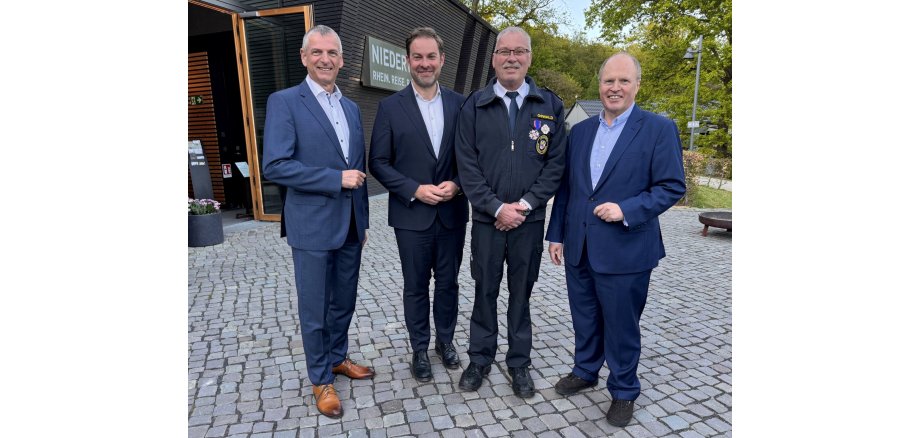 Vier Männer in hellblauem Hemd und dunkelblauem Anzug stehen vor einer Halle, die mit horizontalen Holzbalken verkleidet ist. Der zweite Mann von rechts trägt eine Krawatte, eine Brille und eine Jacke mit seinem Namen und zwei Orden sowie einer Plakette. Der Boden, auf dem sie stehen ist mit Kopfsteinpflaster gepflastert. Im Hintergrund sind grüne Bäume.