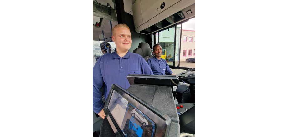 In einem Linienbus sitzen zwei junge Männer in blauem, langärmligem Polo-Shirt. Der Eine sitzt hinter dem Lenkrad und der andere daneben. Im Vordergrund ist das Display der Fahrzeugkamera mit einem Auto zu sehen.