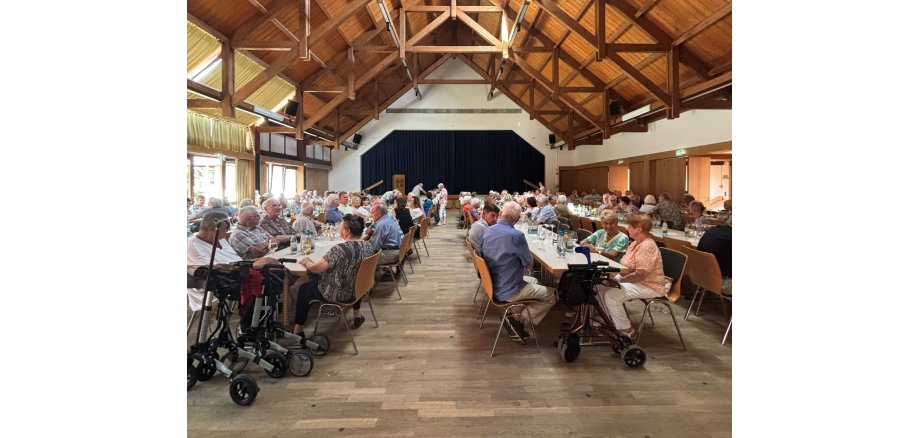 In einem Saal mit Holzgebälg im Dach und einer Bühne am Ende des Raumes stehen mehrere Tischreihen, an denen Personen sitzen. Im Vordergrund stehen mehrere Rollatoren.