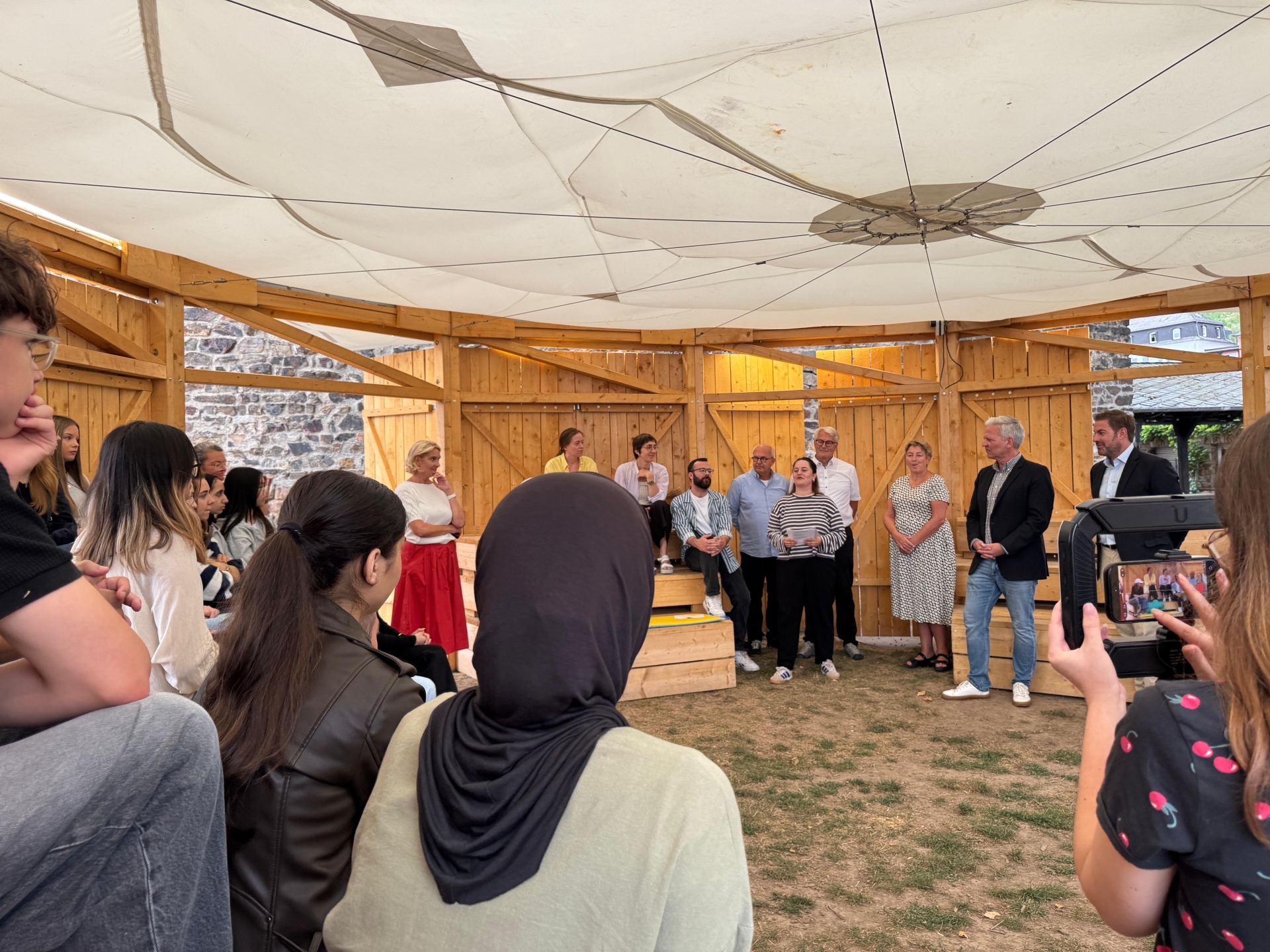 In einem Pavillon aus Holzwänden und einem weißen Tuchdach, sitzen mehrere Personen auf mehreren Holzrängen oder stehen daneben. Der Boden ist eine Grasfläche mit grünem und braunem Gras.