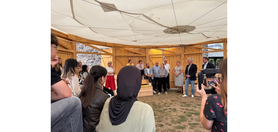 In einem Pavillon aus Holzwänden und einem weißen Tuchdach, sitzen mehrere Personen auf mehreren Holzrängen oder stehen daneben. Der Boden ist eine Grasfläche mit grünem und braunem Gras.