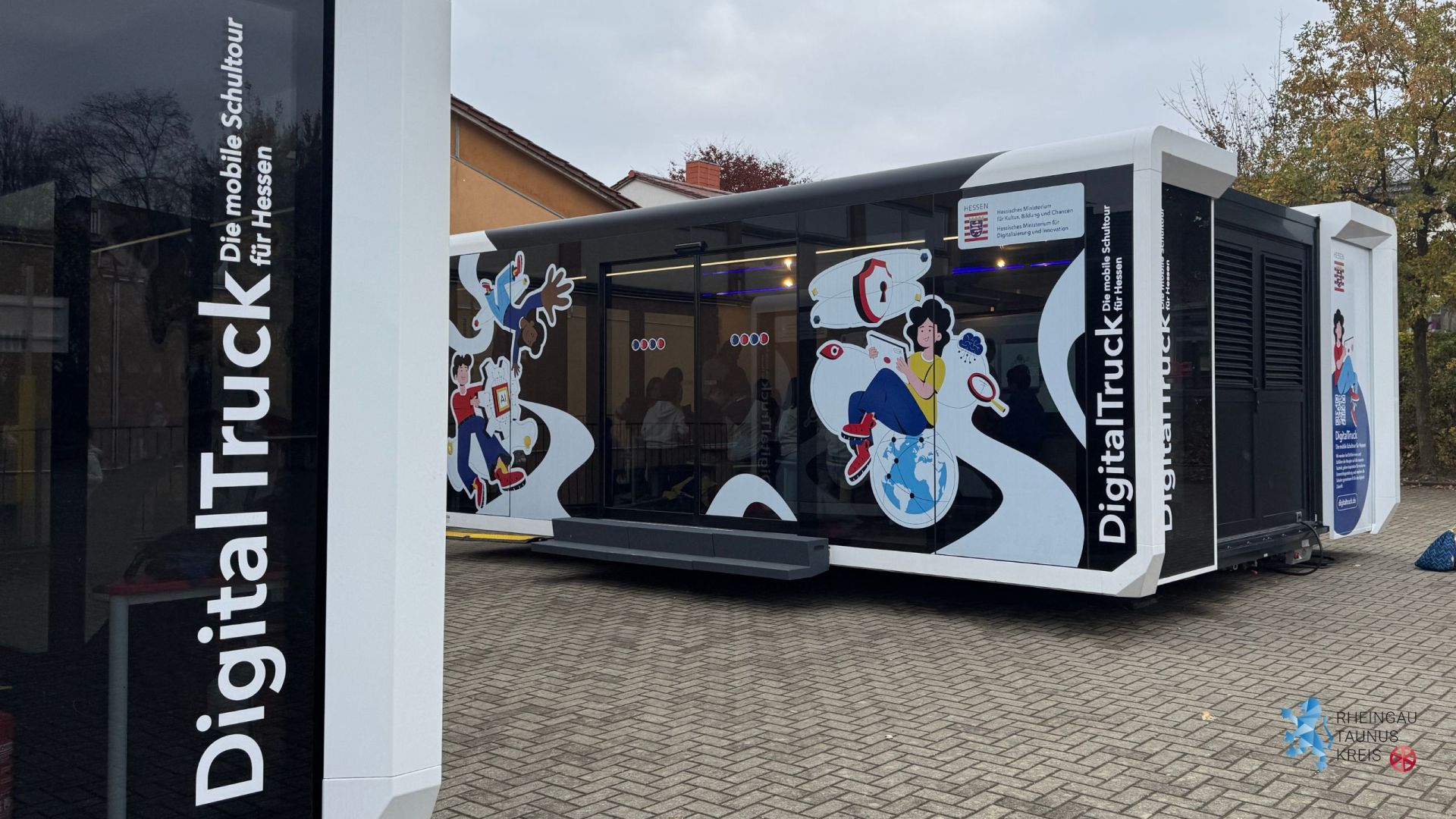 Auf einer gepflasterten Fläche stehen zwei große Container-Anhänger, die mittig ausgefahren wurden. Es sind Glastüren und Glasfenster, die mit gezeichneten Motiven beklebt sind. Unter anderem ist eine Regenwolke, eine Lupe ein Schloss, ein Mädchen mit Tablett in der Hand, die auf einer Erdkugel sitzt und zwei weitere Jugendliche, die Eine scheint schwerelos auf dem Kopf zu schweben.