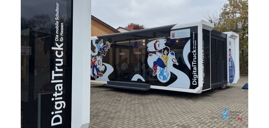 DigitalTruck - 1 Auf einer gepflasterten Fläche stehen zwei große Container-Anhänger, die mittig ausgefahren wurden. Es sind Glastüren und Glasfenster, die mit gezeichneten Motiven beklebt sind. Unter anderem ist eine Regenwolke, eine Lupe ein Schloss, ein Mädchen mit Tablett in der Hand, die auf einer Erdkugel sitzt und zwei weitere Jugendliche, die Eine scheint schwerelos auf dem Kopf zu schweben.