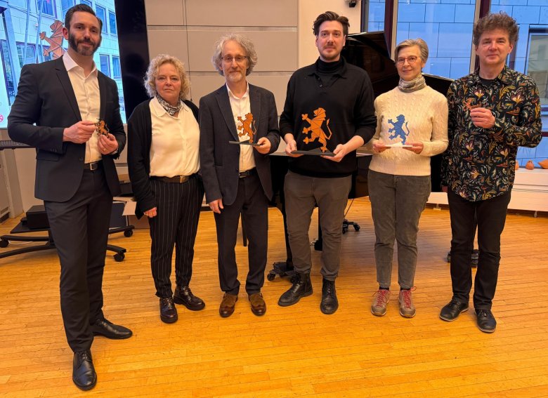 Vier Männer und zwei Frauen  mit Hosen und drei mit weißen Hemden und Blusen und dunklem Sakko und Blazer, einer mit einem floralem Hemd auf schwarzem Untergrund und ein Mann mit schwarzem Pullover und eine Frau mit beigem Pullover stehen vor einer Fensterfront und einem Bildschirm. Vier Personen halten je einen hellbraunen hessischen Löwen mit dunklen Strichen, die Flächen abgrenzen und eine Frau hält den gleichen Löwen, der jedoch die Farbe blau hat.