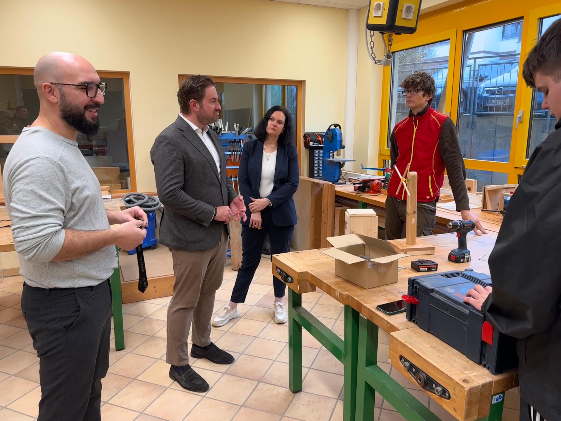 In einem Raum mit Fenstern mit gelben Fensterrahmen, stehen mehrere Werkbänke mit Holzplatten und grünen Metallbeinen. Davor stehen vier Männer und eine Frau.