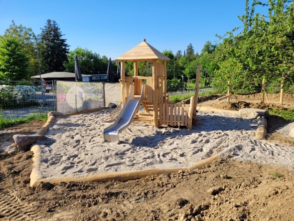 Ein Holzturm mit Dach, einer Holztreppe und einer Rutsch aus Metall sowie ein Holzzaun steht auf einem Platz, der mit Sand gefüllt ist. Umrandet ist er mit Holzbaumstämmen. Außen rum ist Erde und im Hintergrund Holzstämme zum Balancieren und Stangen zum Turnen, verschiedenen Bäume und ein blauer Himmel.