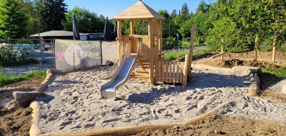Ein Holzturm mit Dach, einer Holztreppe und einer Rutsch aus Metall sowie ein Holzzaun steht auf einem Platz, der mit Sand gefüllt ist. Umrandet ist er mit Holzbaumstämmen. Außen rum ist Erde und im Hintergrund Holzstämme zum Balancieren und Stangen zum Turnen, verschiedenen Bäume und ein blauer Himmel.