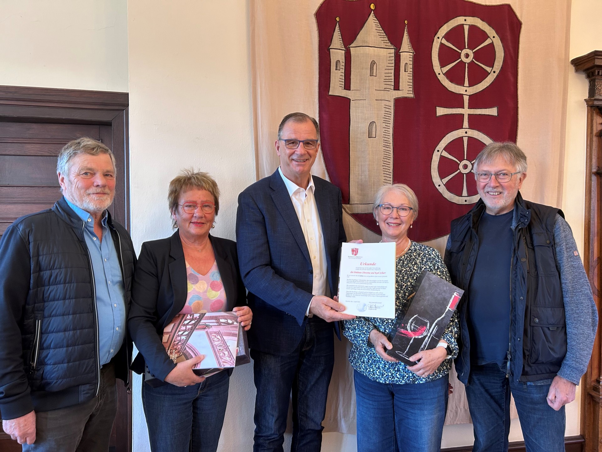 Vor dem Gemeinde-Wappen in weinrot, mit beigem Doppelrad und der Burg stehen drei Männer und zwischen den Männern zwei Frauen. Die linke Frau hat ein Heft über eine Kirche in der Hand und die andere Frau trägt ein Weinpräsent. Der Mann in der Mitte hat eine Urkunde in der Hand.