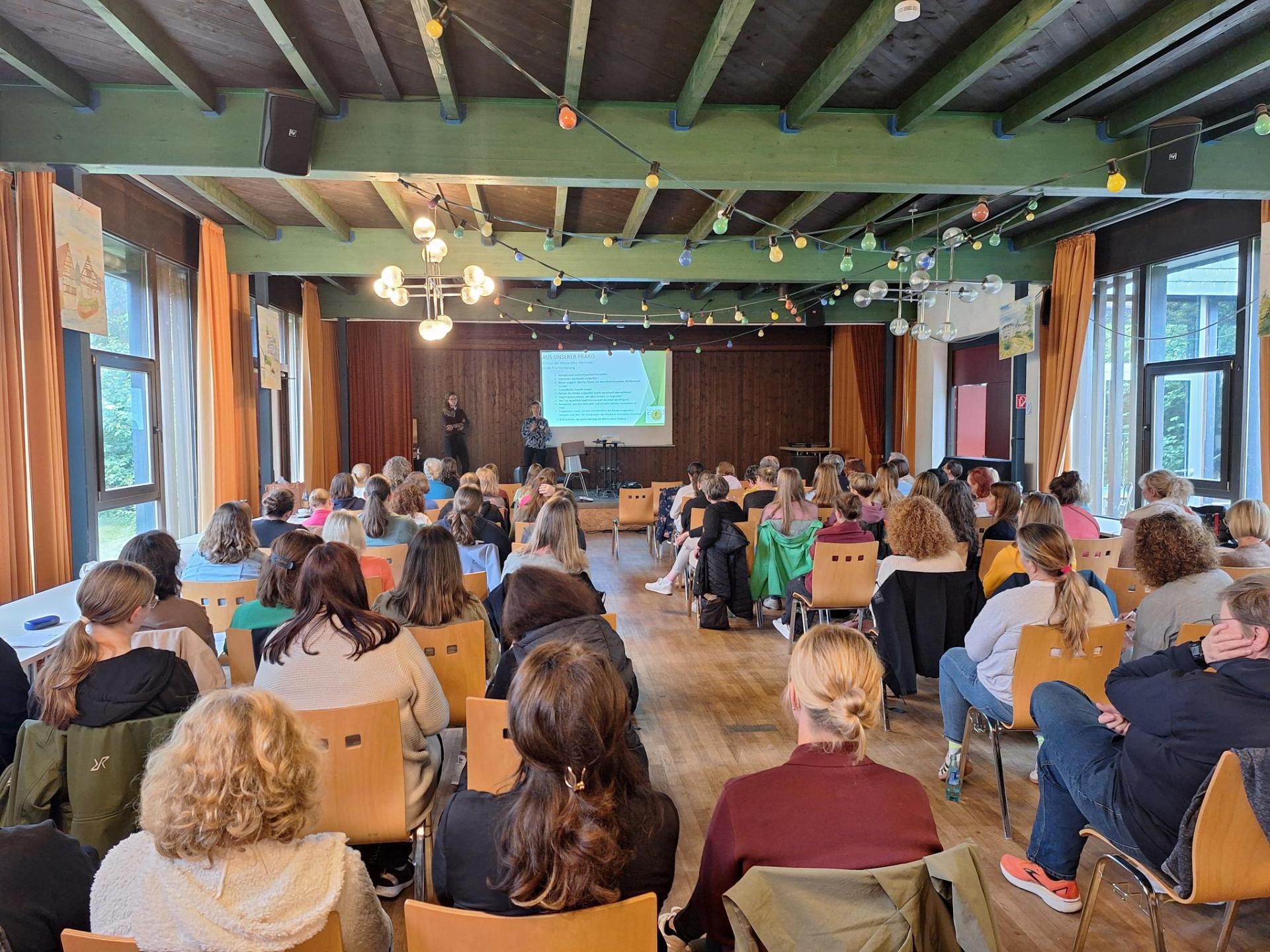Blick von Hinten auf ein Publikum, das auf Holzstühlen sitzt. Vorn ist ein Bühne, auf der zwei Personen neben einer Leinwand stehen.