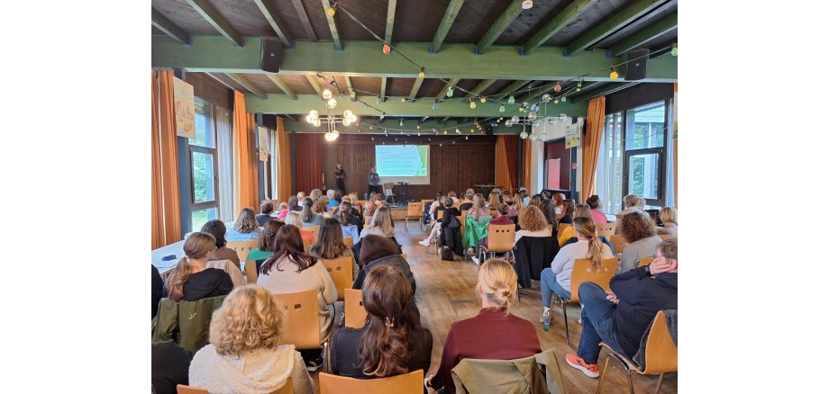 Blick von Hinten auf ein Publikum, das auf Holzstühlen sitzt. Vorn ist ein Bühne, auf der zwei Personen neben einer Leinwand stehen.