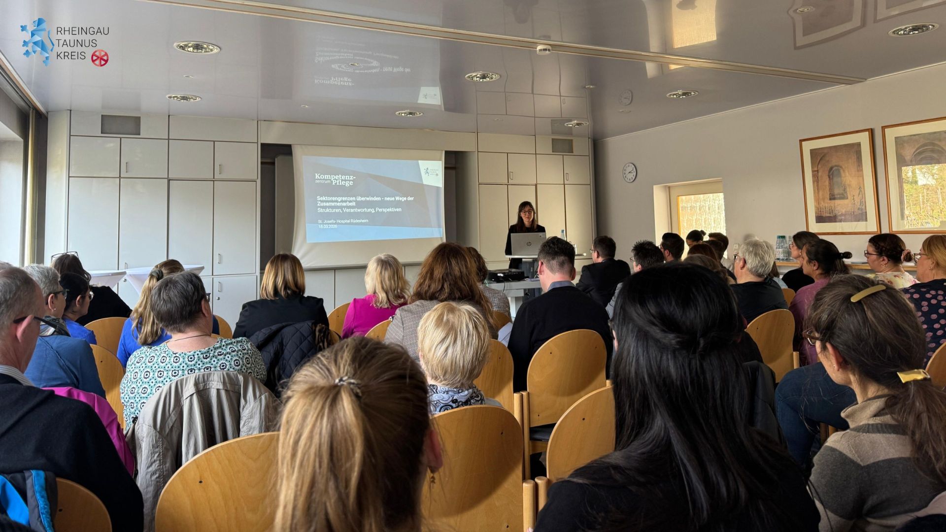 Eine Frau mit Brille und langen Haaren, steht hinter einem Laptop, neben einer Leinwand und einer weißen Schrankwand. Im Raum sitzen Menschen auf braunen Stühlen. An der Wand hängt eine runde Uhr und zwei große Bilder mit bronze-farbenen Rahmen.
