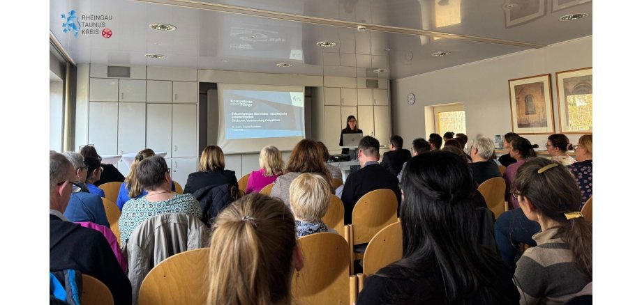 Newsfeed Bild - 260318_Veranstaltung Kompetenzzentrum Pflege Eine Frau mit Brille und langen Haaren, steht hinter einem Laptop, neben einer Leinwand und einer weißen Schrankwand. Im Raum sitzen Menschen auf braunen Stühlen. An der Wand hängt eine runde Uhr und zwei große Bilder mit bronze-farbenen Rahmen.