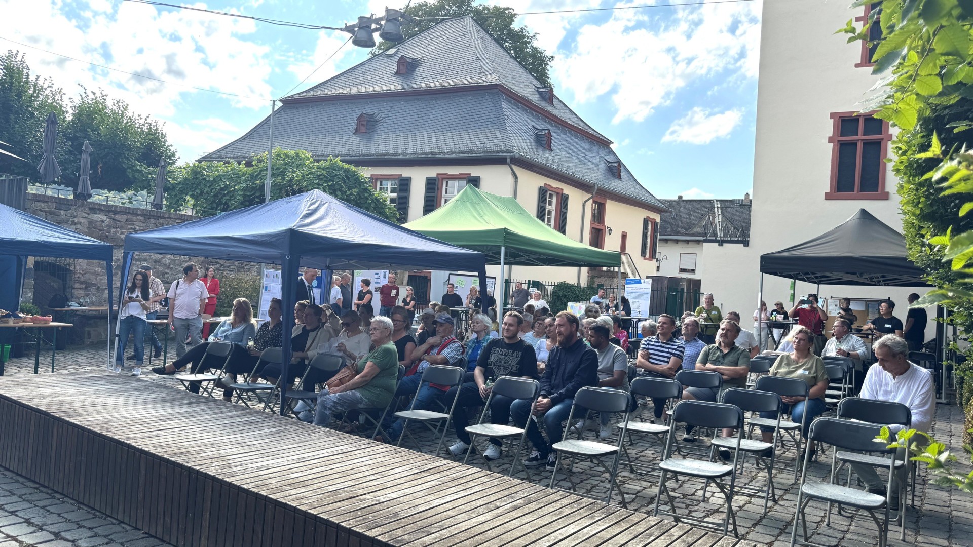 Hinter einer breiten, rechteckigen Holzfläche sind Klappstuhlreihen, auf denen z.T. Menschen sitzen. Es ist ein schwarzer, ein grüner und zwei blaue Pavillons aufgebaut. 
