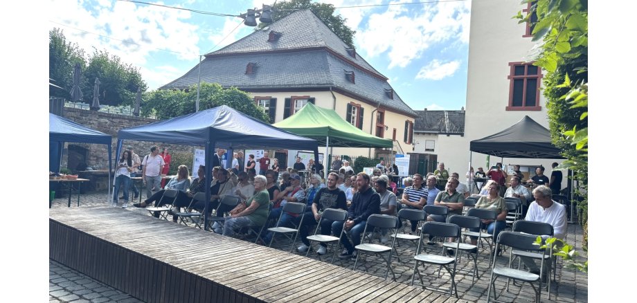 Hinter einer breiten, rechteckigen Holzfläche sind Klappstuhlreihen, auf denen z.T. Menschen sitzen. Es ist ein schwarzer, ein grüner und zwei blaue Pavillons aufgebaut.