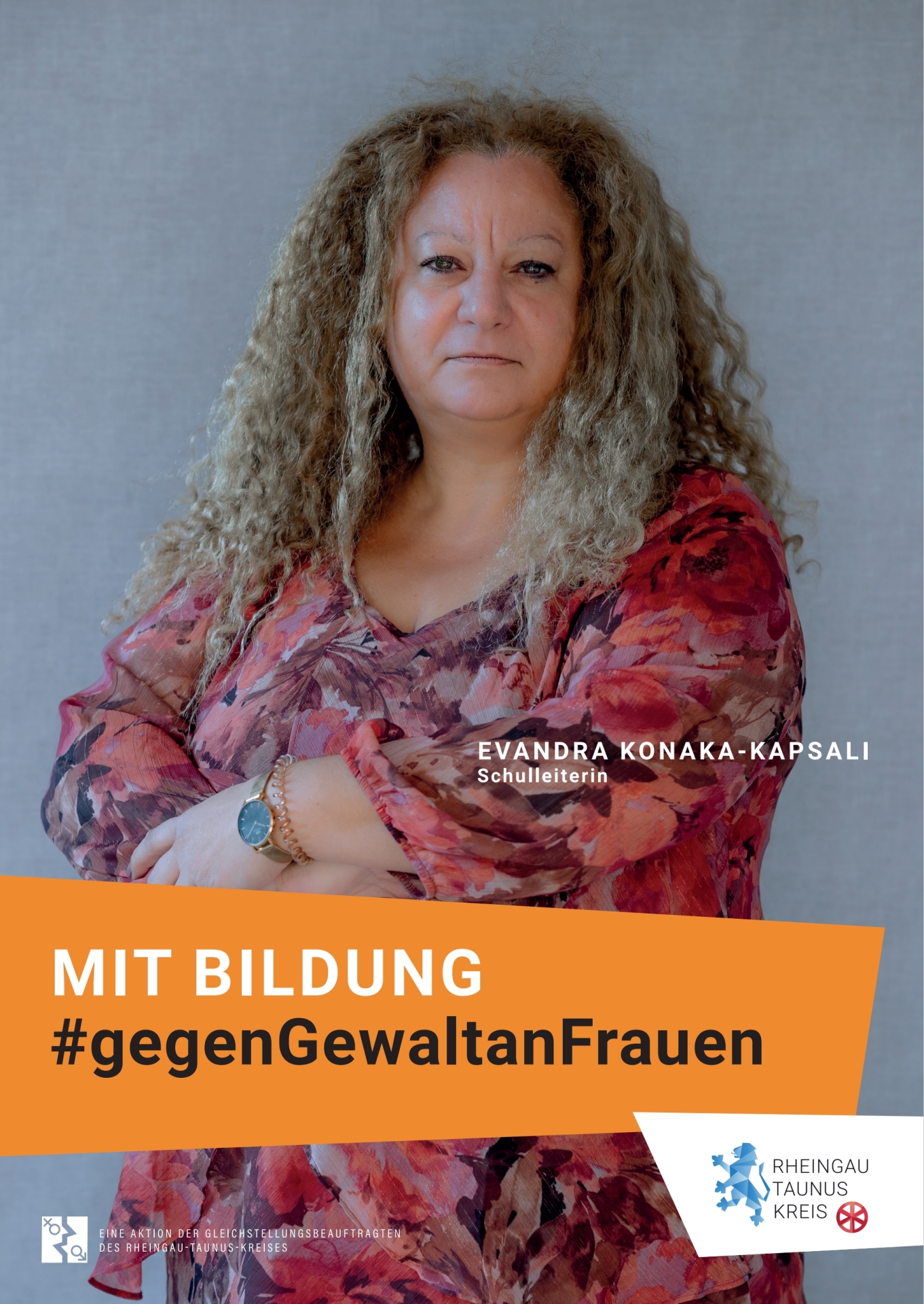 Vor einem grauen Hintergrund steht eine Frau in rosa-rot-weinrot-geblümter Bluse. Sie verschränkt die Arme vor ihren Körper und trägt eine rosefarbene Uhr mit schwarzem Ziffernblatt sowie ein rosa Spiralhaarband. Die blonden Haare sind gelockt und lang. Der Blick ist ernst.