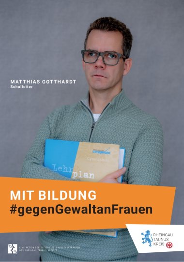 Vor einem grauen Hintergrund steht ein Mann im hellgrünem Strickpulover mit Muster. Er hält einen Ordner mit der Aufschrift "Lehrplan" mit verschränkten Armen vor seinen Körper hält. Er trägt eine grau melierte rechteckige Brille und gruße braune Haare. Der Blick ist ernst.