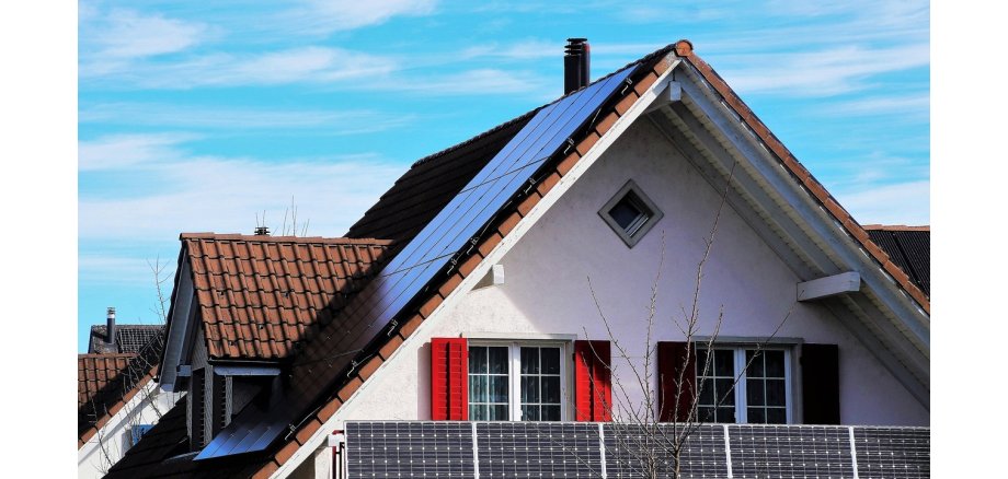 Man sieht das Dachgeschoss mit einem Dachgiebel. Hinter dem Balkon mit fünf Solarpanelen sind zwei Paar Sprossenfenster mit roten Rolläden. Das Haus ist weiß und die Dachziegel rot-braun. Auf dem Dach sind ebenso Solarpanelen.