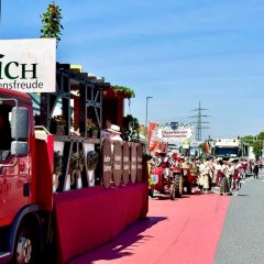 Ein roter Laster mit Holzbalken, runden Schildern und Blumen geschmückt fährt auf einer Straße. Hinten dran steht ein geschmückter Traktor mit Schild und ein weiterer Laster. Es stehen Menschen am Straßenrand und Menschen im Dirndl auf der Straße.