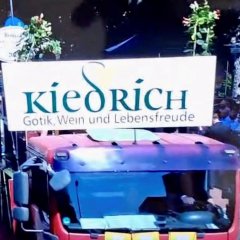 Man sieht den oberen Teil des Führerhauses eines roten Lasters, auf dem ein großes weißes Schild mit dem Namen "Kiedrich, Gotik, Wein und Lebensfreude" steht.