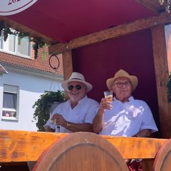 Zwei Männer mit weißem, kurzärmeligem Hemd und Sonnenhut und einem Becher mit Wein in der Hand, sitzen auf einem offenem Wagen, der mit Holz und Stoff geschmückt ist und an dem Trauben und Efeu hängen.