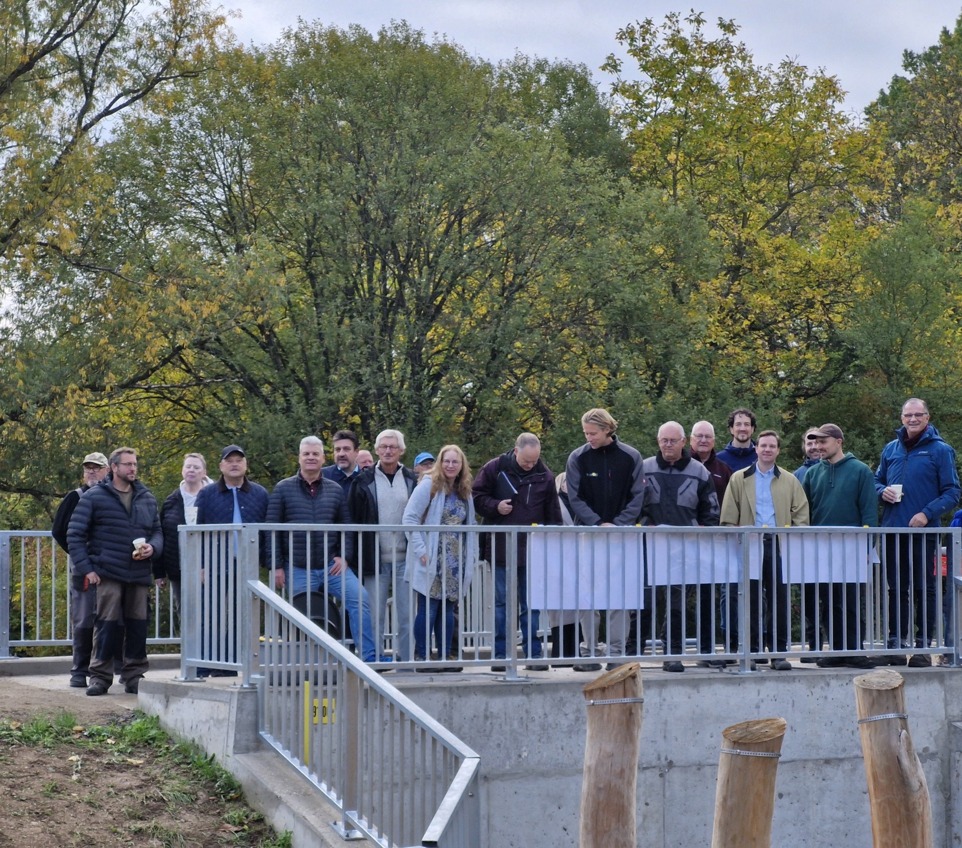 Auf einer Brücke aus Beton und Eisengeländer, stehen 20 Männer und Frauen mit Jacken. Im Hintergrund sind große belaubte Bäume.