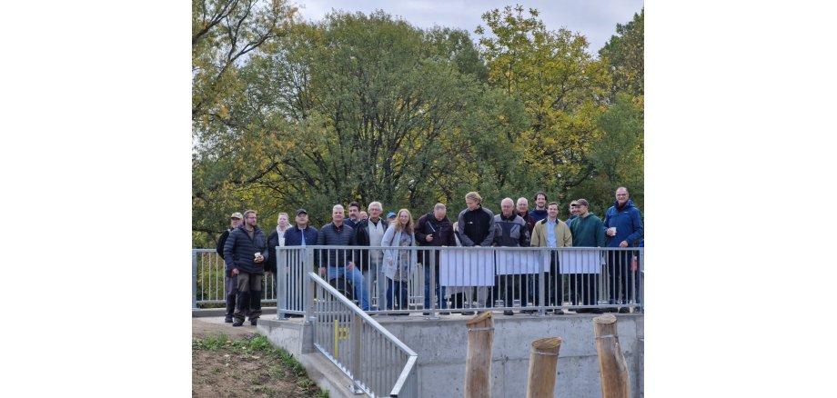 Auf einer Brücke aus Beton und Eisengeländer, stehen 20 Männer und Frauen mit Jacken. Im Hintergrund sind große belaubte Bäume.