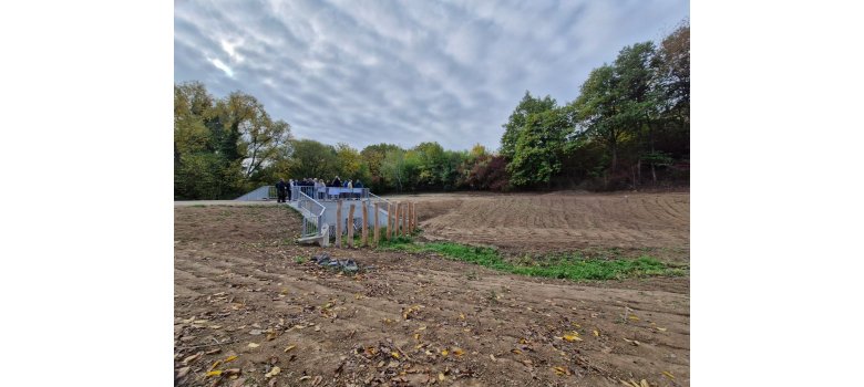 Hochwasserrückhaltebecken "Grünbach" Eine Brücke aus Beton und Metallgeländer stehen mehrere Personen. Vor der Brücke sind 9 große Holzpfeiler und eine große Erdfläche. Im HIntergrund sind belaubte Bäume, ein bewölkter Himmel.