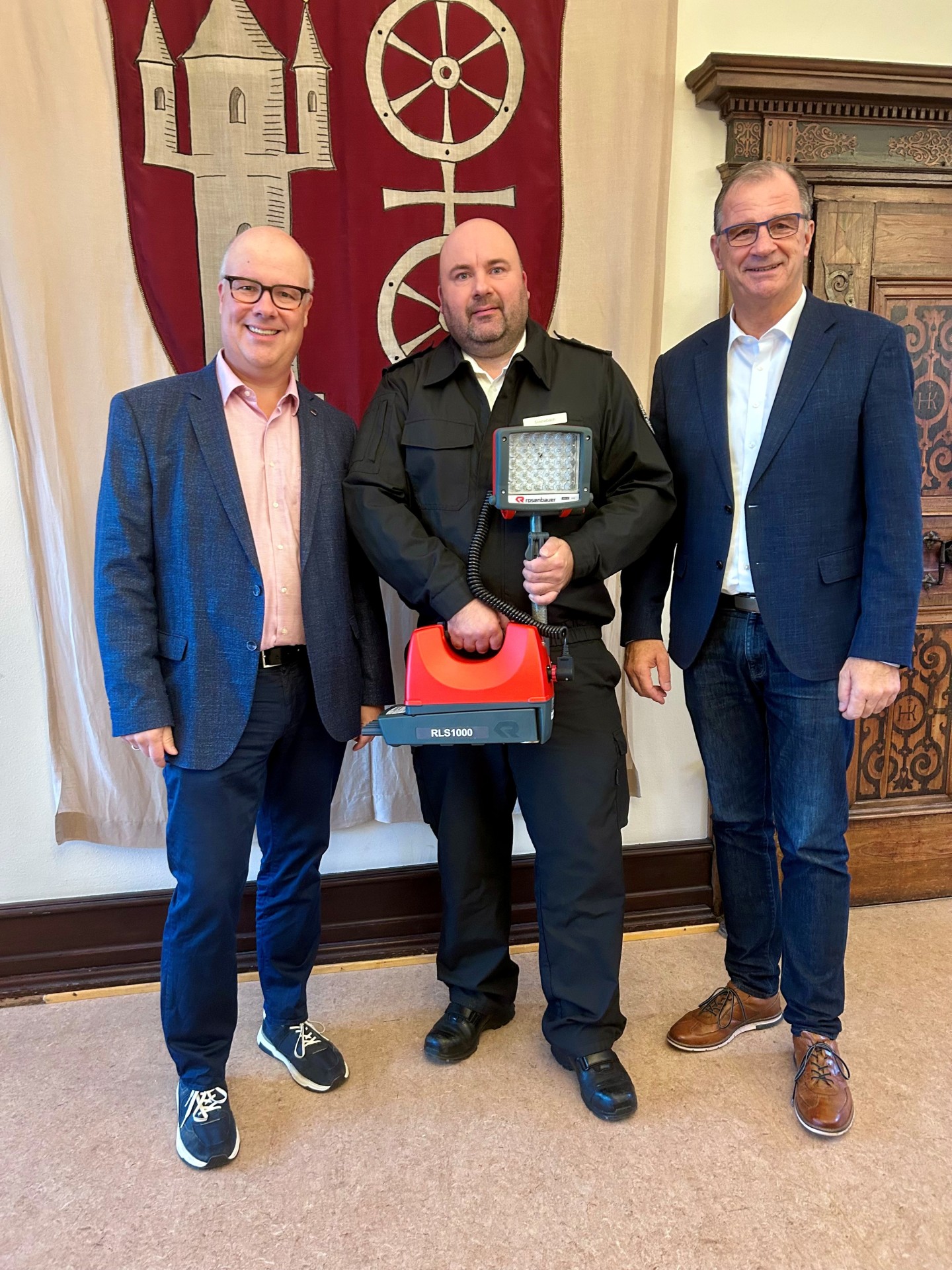 Vor einem großen Wandbild aus Stoff, auf der ein weinrotes Wappen mit einem beigen Doppelrad und einer beigen Burg, stehen drei Männer. Der Mann in der mitte hält ein LED-Beleuchtungssystem in orange und grau in beiden Händen.