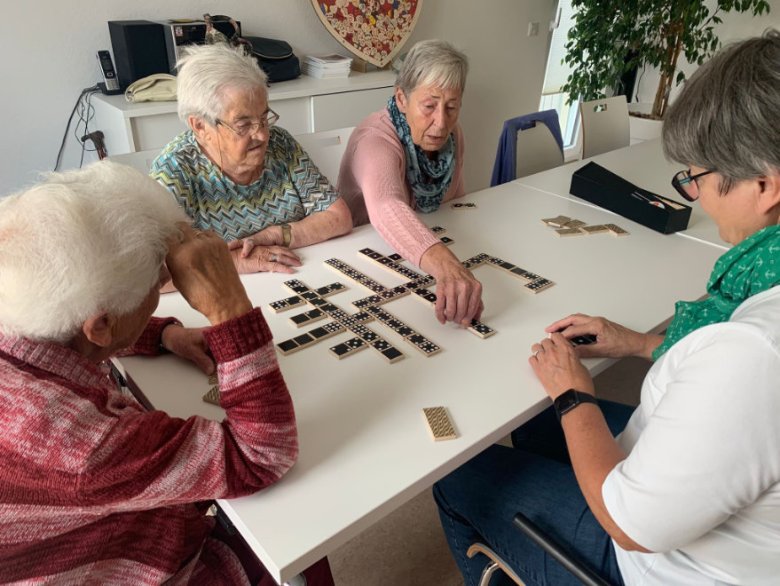 An einem rechteckigen Tisch mit weißer Tischplatte sitzen vier ältere Frauen und Spielen Domino. Die Steine sind schwarz mit weißen Punkten.