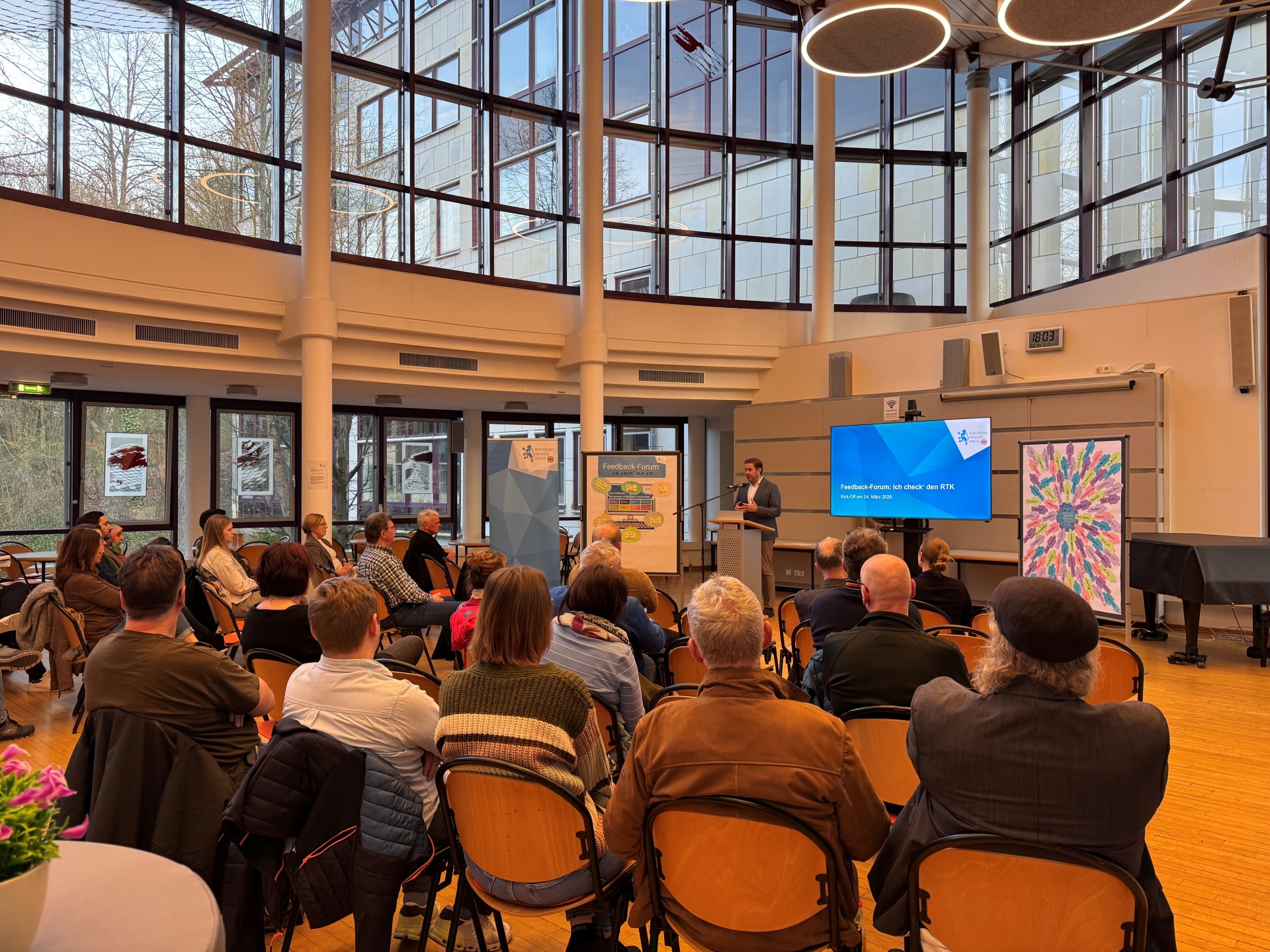 In einem Saal mit einer großen halbrunden Fensterfront sitzen mehrere Personen auf braunen Holzstühlen. Vorn steht an einem Rednerpult ein Mann im Anzug. Neben ihm sind mehrere Leinwände und ein zugedeckter Flügel.