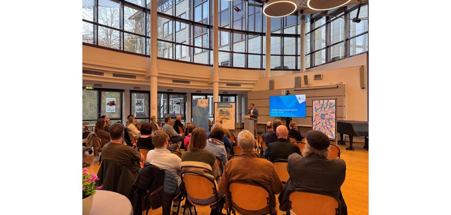 In einem Saal mit einer großen halbrunden Fensterfront sitzen mehrere Personen auf braunen Holzstühlen. Vorn steht an einem Rednerpult ein Mann im Anzug. Neben ihm sind mehrere Leinwände und ein zugedeckter Flügel.