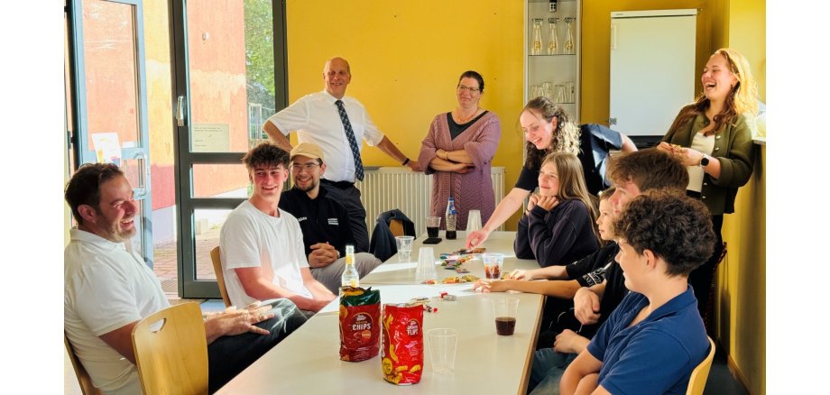 Ein Raum mit gelben Wänden, einer Glasvitrine  mit Gläsern, ein Flipchart und eine geöffnete Tür aus Glas und Metall. An einem weißem Tisch sitzen mehrere Personen, lachend auf Stühlen. Weitere Personen stehen lachend um den Tisch. Auf dem Tisch ist eine geöffnete Chips und Flips-Tüte, Bonbons, Gummibären und durchsichtige Plastikbecher, die teilweise mit Getränken gefüllt sind.